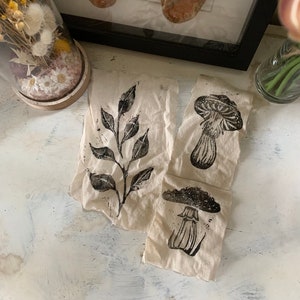 May include: Three fabric squares with black ink block prints of a leafy branch, a mushroom, and a mushroom with a stem. The fabric is a natural beige colour.