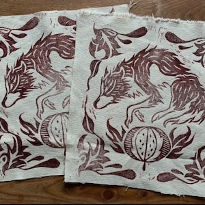 Könnte beinhalten: Zwei braune Stoffquadrate mit einem braunen Wolfsmuster und einem braunen Fruchtmuster. Der Wolf springt über die Frucht. Der Stoff ist hellbeige.