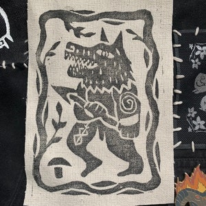 May include: An embroidered patch featuring a stylised wolf with a snarling expression, in black and white. The wolf is surrounded by a decorative frame with a floral design.