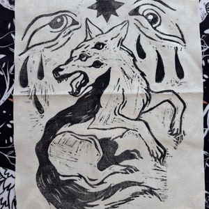 Peut inclure: Impression linogravure en noir et blanc sur tissu représentant un loup à trois yeux, une étoile et deux yeux qui pleurent. Le loup grogne et a une longue queue.