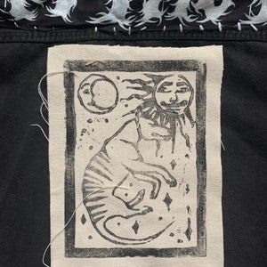 May include: Black and white linocut print of a creature with a sun-like face, surrounded by stars and a crescent moon. The print is framed with a black border and sewn onto a black fabric.