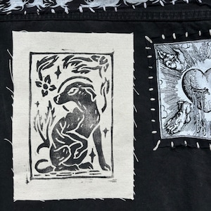 May include: Close-up of a black fabric with two rectangular patches. The left patch features a black and white linocut print of a dog surrounded by floral and star elements. The right patch has a black and white illustration of a heart and hand.