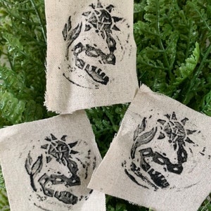 May include: Three small, square, beige fabric patches with black ink printed designs of a stylised sun and a creature. The designs are centred on each patch.