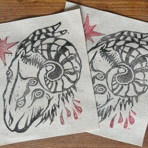 Peut inclure: Deux carrés de tissu imprimés en noir et blanc avec un motif de tête de bélier stylisé. Chaque carré a une étoile rouge dans le coin supérieur et des gouttes de peinture rouge en bas.