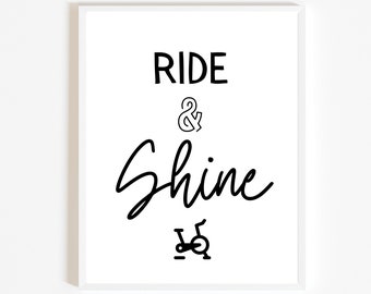 May include: White framed print with the words "RIDE & Shine" in black script and block letters. A black stationary bike icon is at the bottom of the print. The print is a motivational piece for a home gym or fitness space.