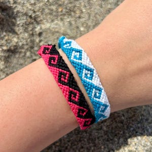 May include: Two handmade friendship bracelets. One bracelet is pink with black geometric patterns. The other bracelet is blue with white geometric patterns. The bracelets are worn on a wrist.