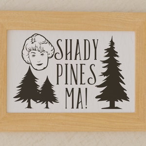 Puede incluir: Una impresión enmarcada con una ilustración en blanco y negro de la cara de una mujer y dos pinos. El texto "Shady Pines Ma!" está escrito en una fuente cursiva.