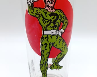 May include: A clear glass tumbler featuring a graphic of a character in a green suit covered in black question marks, standing in front of a large red circle. The character wears a white belt and gloves. The word RIDDLER is printed in red at the base.