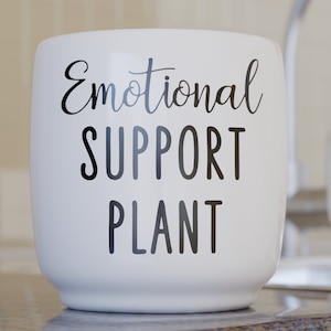 May include: White ceramic plant pot with the words "Emotional SUPPORT PLANT" in black script. The pot is cylindrical and sits on a countertop, with a blurred background of a kitchen.