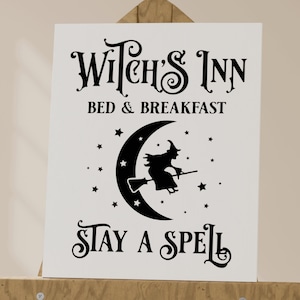 May include: A white sign with black text and a silhouette of a witch flying on a broomstick in front of a crescent moon. The sign reads "Witch's Inn Bed & Breakfast Stay A Spell".