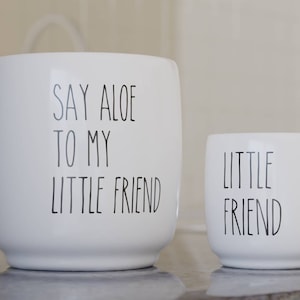 May include: Two white ceramic mugs with black lettering. The larger mug says "Say Aloe to my Little Friend". The smaller mug says "Little Friend".