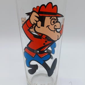 May include: A clear glass tumbler featuring a colorful graphic of the cartoon character Dudley Do-Right wearing a red uniform and hat with blue trousers. The character is depicted in a running pose. The text Dudley Do-Right is printed at the base.