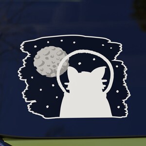 May include: A white silhouette of a cat wearing a space helmet, looking up at a crescent moon with craters, against a dark blue background with white stars.