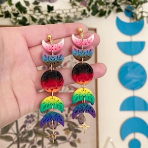 May include: A pair of long earrings with a moon phase design. The earrings display a rainbow of colours, including pink, blue, green, yellow, and purple. Each phase is decorated with leaf patterns and a gold star charm.