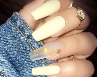 May include: Close-up of a hand with long, yellow nails. Some of the nails have clear polish with gold butterfly decals. The hand is wearing gold rings.