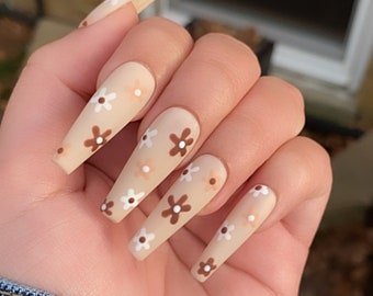 May include: A set of long, nude-colored press-on nails with a floral design. The flowers are white with brown centers and are scattered across the nails.