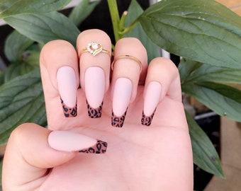 May include: Close-up of a hand with long, nude-colored, almond-shaped nails. The tips of the nails are painted with a leopard print design.