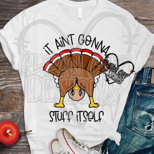Op de afbeelding: Wit T-shirt met een cartoon-illustratie van een kalkoen en de tekst "It ain't gonna stuff itself".