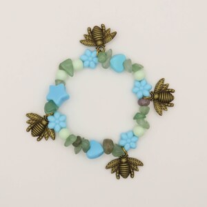 Puede incluir: Un brazalete de cuentas azul claro y verde con abalorios de abeja y detalles de flores.
