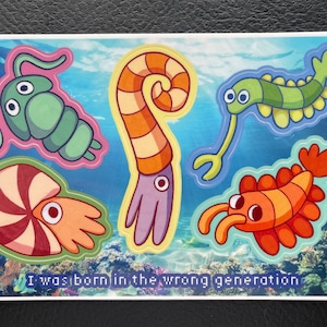 May include: A sticker sheet featuring five colourful cartoon sea creatures, including a shrimp, a nautilus, a squid, a worm, and a trilobite. The sticker sheet has a blue background with a text overlay that reads "I was born in the wrong generation."