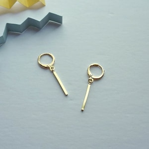 May include: A pair of gold earrings with a thin, straight bar hanging from a small hoop. The earrings are on a white surface.