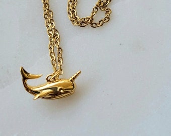 May include: A gold chain necklace with a gold narwhal pendant. The narwhal is facing to the right and has a long, pointed horn.