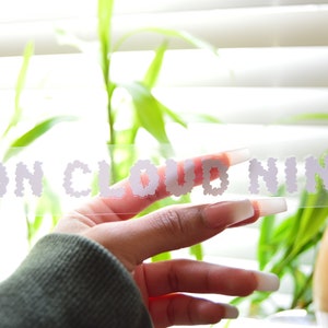 May include: A clear sticker with the text "ON CLOUD NINE" in white, made of small cloud shapes. The sticker is being held by a hand with long, white nails.