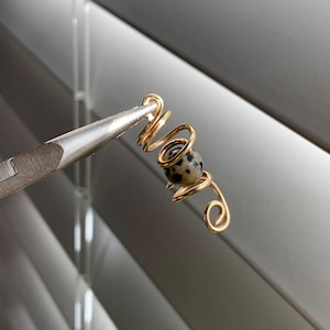 Puede incluir: Un pendiente de oreja dorado con un diseño en espiral, con una pequeña cuenta moteada. El pendiente está sujeto por unos alicates plateados sobre un fondo borroso de persianas.