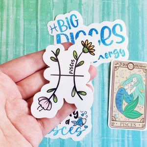 May include: A set of five stickers featuring a mermaid, a flower with a stem, and the zodiac sign Pisces. The stickers are white with blue, green, and pink accents. The text on the stickers reads "Big Pisces Energy" and "Pisces".