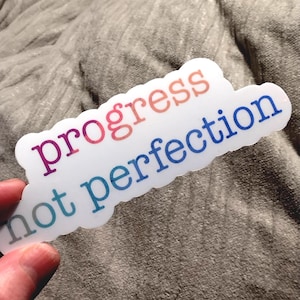 May include: A white sticker with the words "progress not perfection" in gradient colors of pink, orange, and blue. The sticker is held by a hand against a gray, textured background.