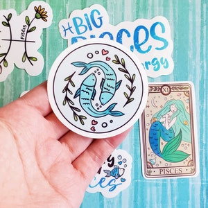 May include: A set of five stickers featuring the astrological sign Pisces. The stickers are round and feature a variety of designs, including two fish swimming, a mermaid, and the word "Pisces".