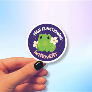 May include: A purple circle sticker with a green frog and white flowers. The text on the sticker reads "High Functioning Introvert".