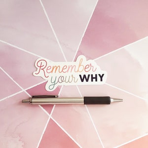 Puede incluir: Una pegatina blanca con el texto "Remember your WHY" en colores arco&iacute;ris sobre un fondo geom&eacute;trico rosa y blanco con un bol&iacute;grafo plateado.