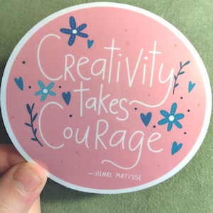 May include: A round sticker with a pink background and white border. The sticker features the words "Creativity Takes Courage" in white script, with blue floral and heart accents. The quote is attributed to Henri Matisse.