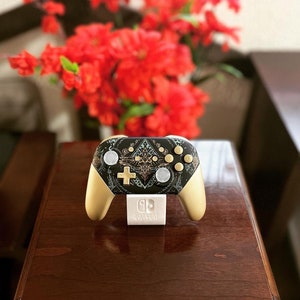 May include: A gold Nintendo Switch controller with a black and white design featuring a stylized bird and geometric patterns. The controller is on a white stand with the Nintendo Switch logo.