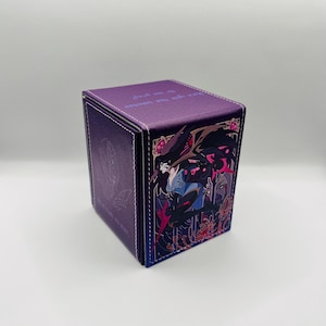 May include: A purple card storage box with a detailed illustration of a character with long black hair and a blue and black outfit. The box has white stitching and text on the top.