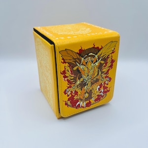 May include: Yellow card storage box with a detailed illustration of a winged dragon with red and orange flames. The box has a square shape and a black interior. The top and sides of the box have a subtle embossed pattern.