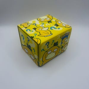 May include: A yellow box featuring a repeating pattern of a cartoon duck with question marks. The box has a cube shape and the word "Pokemon" is visible on one side. The duck is yellow with a white belly and a confused expression.