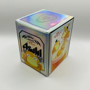 May include: A holographic, square box featuring a cartoon Psyduck character and the Asahi beer logo. The box has a silver trim and a reflective, iridescent surface. The design includes the text "Asahi" and "Psy".