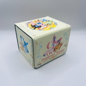 May include: A cream-colored, cube-shaped storage box featuring colorful cartoon characters. The box has a circular design on the top and the words "Pokemon Eevee" on the side. The box is decorated with various cartoon characters and floral accents.