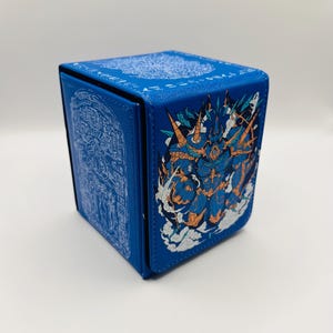 May include: A blue, square card storage box with detailed artwork. The box features a stylized illustration of a creature with orange and blue accents. The box also has a detailed white design on the top and side.