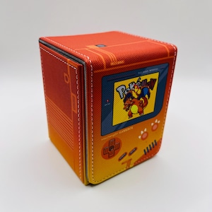May include: A square, orange card storage box with a retro video game design. The box features a screen with a Pokémon graphic and the word "Pokémon" in yellow. The box has a stitched border and a magnetic closure.
