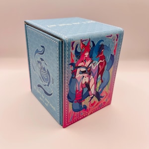 May include: A light blue, square card storage box with a colorful illustration of a character with fox ears and tails. The box has a stitched border and a design on the side.