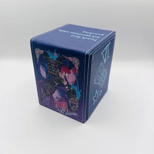 May include: A blue rectangular deck box with white stitching, featuring a graphic of a character with red hair and large gauntlets. The top surface displays the text "Punch first, ask questions while punching" in white, with additional symbols on the side.
