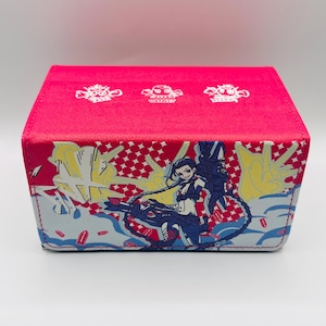 May include: A rectangular, pink storage box with a colorful graphic of a character with a large gun. The box has a red top with white symbols. The sides feature a comic-style design with blue, yellow, and red accents.