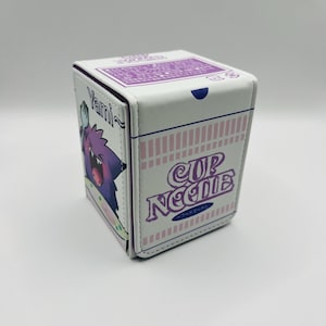 May include: White, cube-shaped storage box with a purple and pink Cup Noodles design. The box features a cartoon image of a purple ghost-like character and the word "Yami" on one side. The top has a small blue clasp.