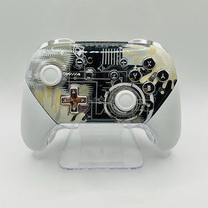 May include: A white and black Nintendo Switch controller with a custom design featuring a circuit board pattern and gold accents.