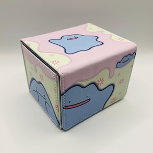 May include: A square, hinged box featuring a cartoon character design. The box has a pink top with a blue character and a light green background with a repeating floral pattern. The sides of the box also feature the blue character.