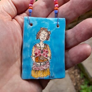 May include: A hand-painted ceramic pendant featuring a woman holding a small animal against a blue backdrop. The pendant has a beaded handle with colourful beads. The woman wears a pink and yellow dress. The pendant is rectangular.