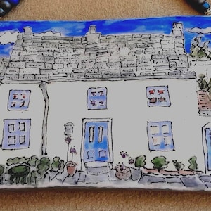 May include: Watercolour painting of a white house with a grey stone roof and blue accents. The house features multiple windows and a blue front door. The painting includes a blue sky and decorative plants.